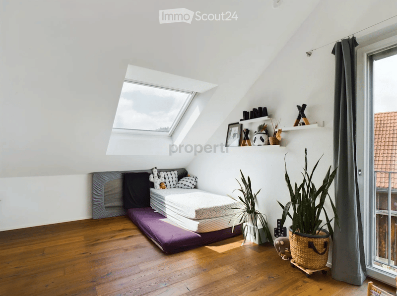 Cozy attic bedroom with wooden flooring, slanted white ceilings, skylight window, and a bed with pillows. Shelves hold decorative items and plants, creating a warm and inviting atmosphere.