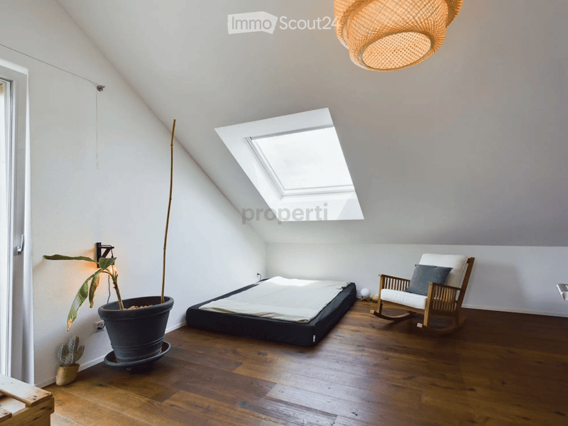 Bright, airy bedroom with wooden flooring, slanted ceiling, skylight window, bed, rocking chair, and potted plants.