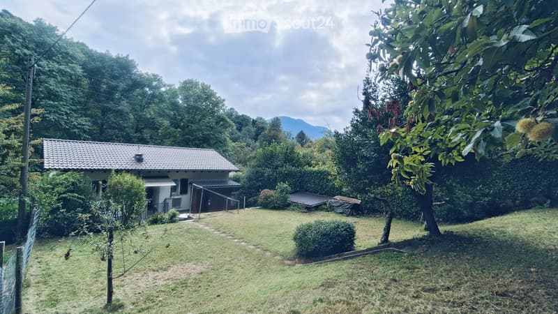 A small, single-story house with a tiled roof, surrounded by a well-maintained lawn, trees, and shrubs. The house has a covered entrance and is situated in a lush, green area with mountains in the background.