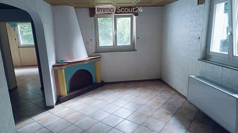 Empty room with tiled floor, white textured walls, and a decorative fireplace with mosaic tiles. Windows provide natural light, and there is a radiator on the right side.
