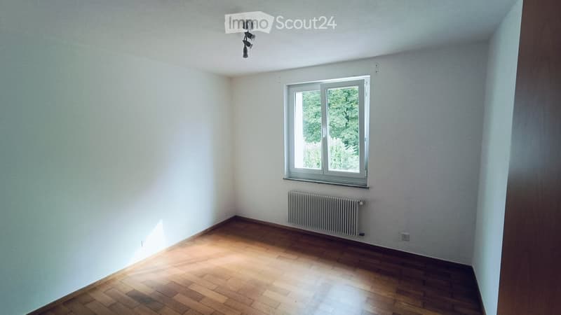 Empty room with wooden flooring, white walls, a single window with a view of greenery, and a radiator below the window.