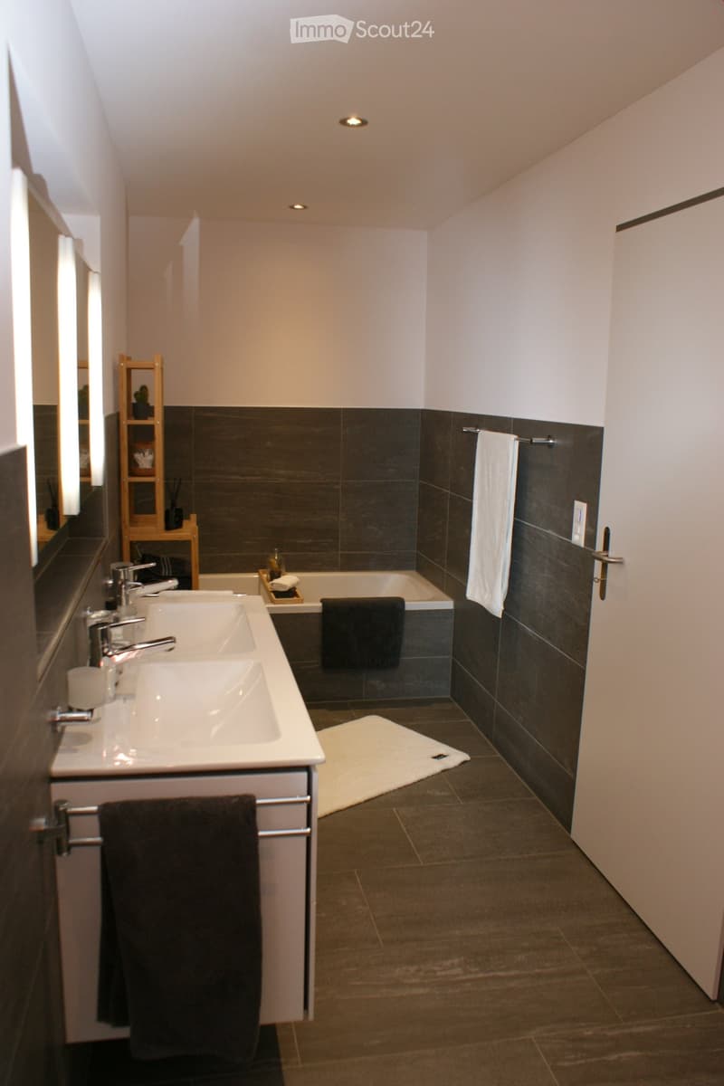 Modern bathroom with double sink, bathtub, dark tiled walls, and a small wooden shelf with decorative items.