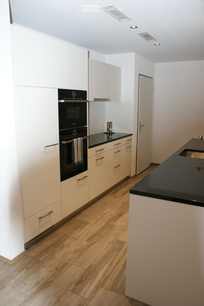 Modern kitchen with white cabinets, black countertops, wooden flooring, and stainless steel appliances including an oven and sink.