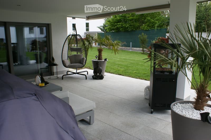 A modern outdoor patio with a wicker hanging chair, a small table, a lounge chair, a Buddha statue, a portable fireplace, and several potted plants, including palm trees and decorative containers.