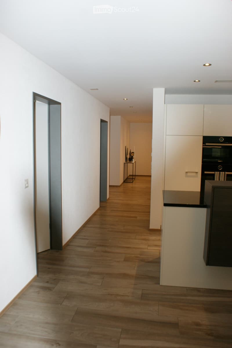 A modern, well-lit hallway with wooden flooring, white walls, and recessed ceiling lights. The hallway leads to a kitchen area with white cabinets and a black countertop.