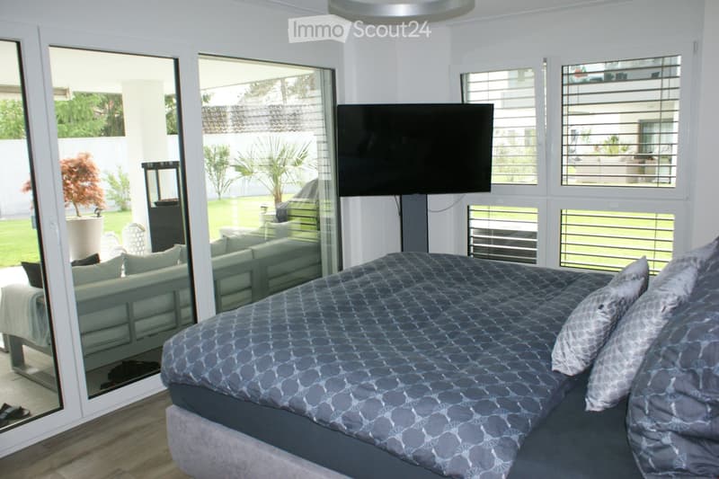Modern bedroom with a large bed, grey bedding, a sofa, and a flat-screen TV mounted on the wall. Large windows and glass doors lead to a garden area.