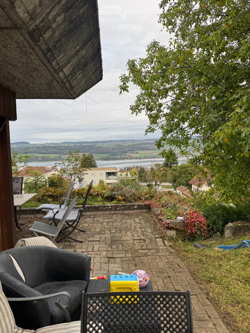A spacious balcony with a brick floor, overlooking a scenic landscape with a river and distant hills. Features a black leather sofa, a small table with colorful items, and several chairs.