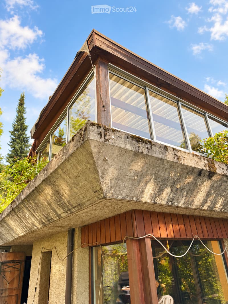 Modern house with a unique, angular design featuring a combination of concrete and wooden elements. The roof has a distinctive, sloped structure with large windows, and the facade includes a mix of textured concrete and wooden panels.