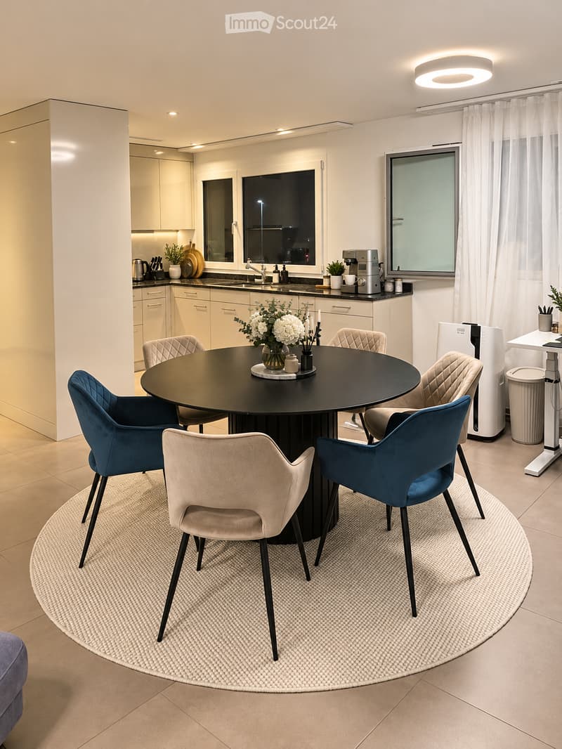 Modern dining area with a round black table surrounded by six chairs, including blue and beige upholstered seats. A flower vase with white flowers is placed at the center of the table. The area is adjacent to a sleek kitchen with white cabinets, a sink, a