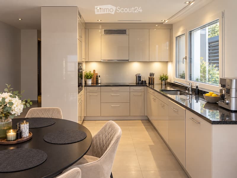 Modern kitchen with white cabinets, black countertops, and a round dining table with chairs. Features include a coffee maker, sink, and decorative items like a vase with flowers and candles.