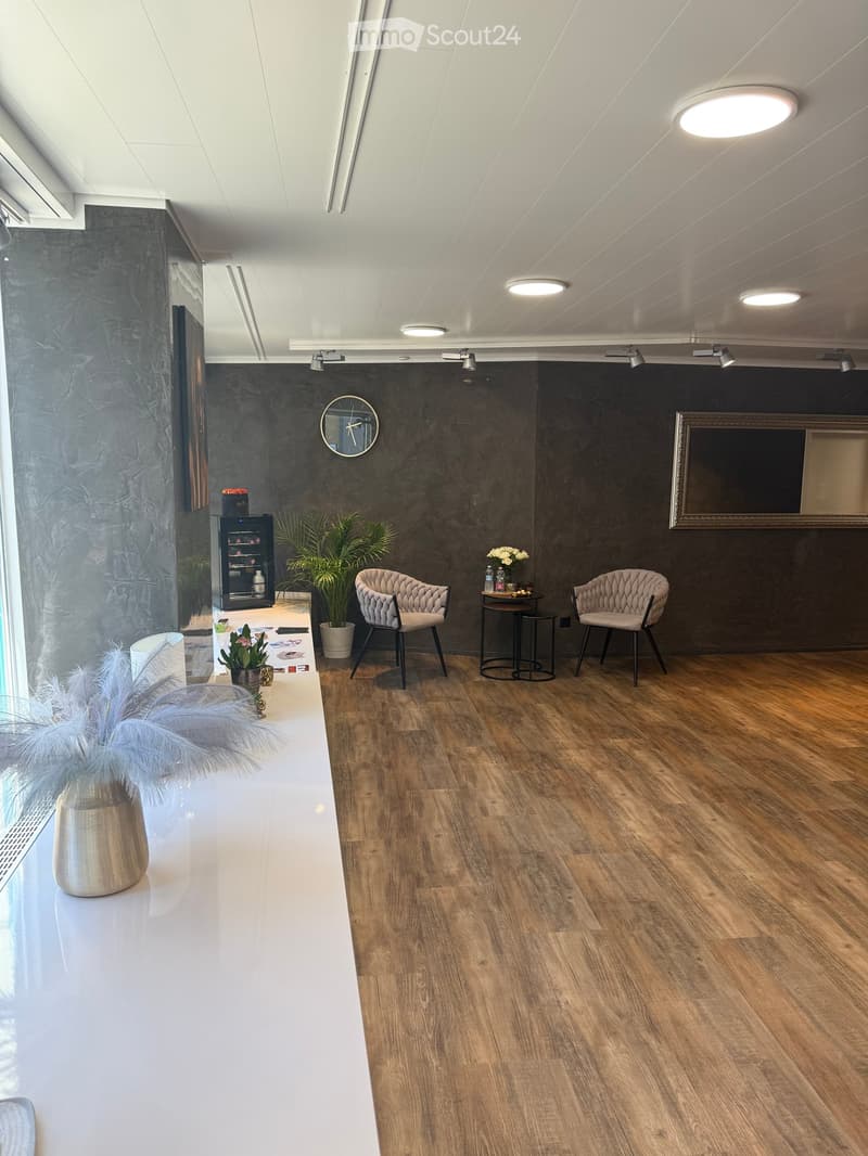 Modern, well-lit room with wooden flooring, two upholstered chairs, a small table, a refrigerator, a mirror, and decorative plants.