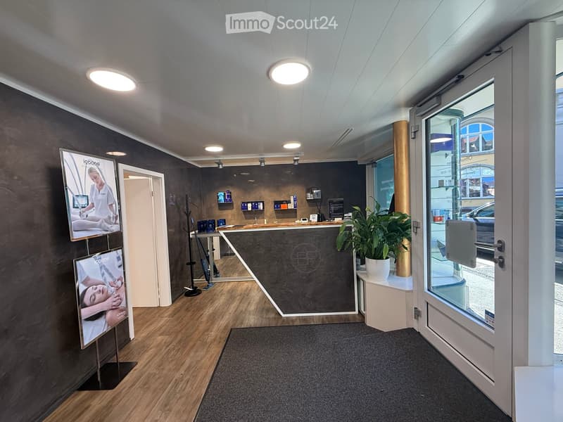 Modern reception area with a sleek reception desk, a large screen displaying images, shelves with supplies, a potted plant, and cleaning tools near the entrance.