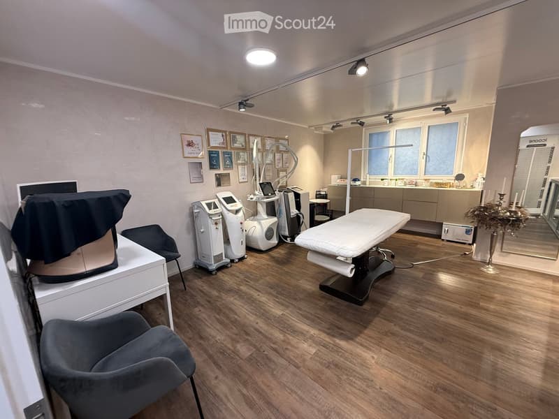 A modern, well-lit room with wooden flooring, a medical examination bed, a large mirror, a cabinet, a chair, and several medical devices.