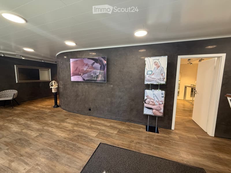 Modern waiting area with wooden flooring, dark textured wall, a sofa, a screen, posters, and a mirror.