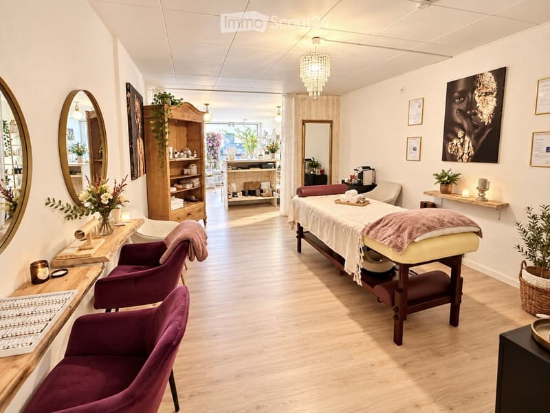 A well-lit, modern spa or relaxation room with wooden flooring, a massage table, two purple armchairs, a large mirror, a wooden cabinet, and decorative plants.