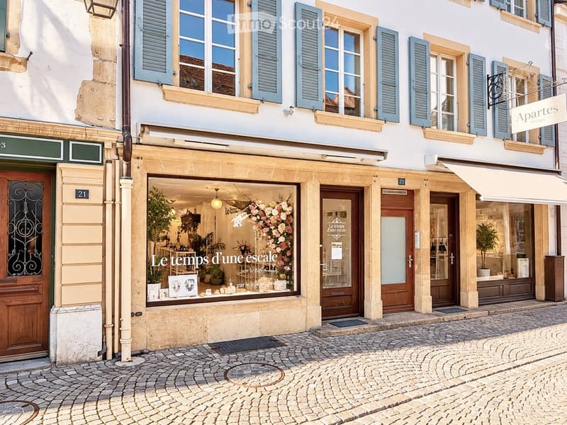 A charming multi-story building with classic European architecture, featuring light-colored stone facades, blue shutters, and multiple storefronts including a flower shop with visible floral arrangements and decorative items inside.