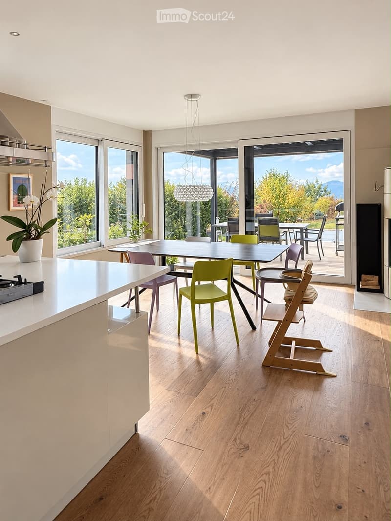 Bright, modern kitchen and dining area with light wood flooring, white cabinetry, and large windows overlooking a scenic outdoor space. Features a central dining table with colorful chairs, a high chair, and a kitchen island with a gas stove.