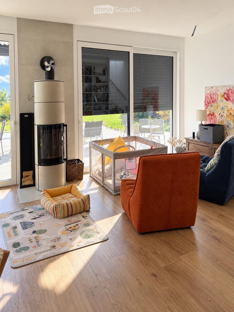 Bright living room with wooden flooring, large windows offering a view of the garden, a modern fireplace, a cozy orange armchair, a blue armchair, a dog bed, a playpen, and a small table with chairs in the background.