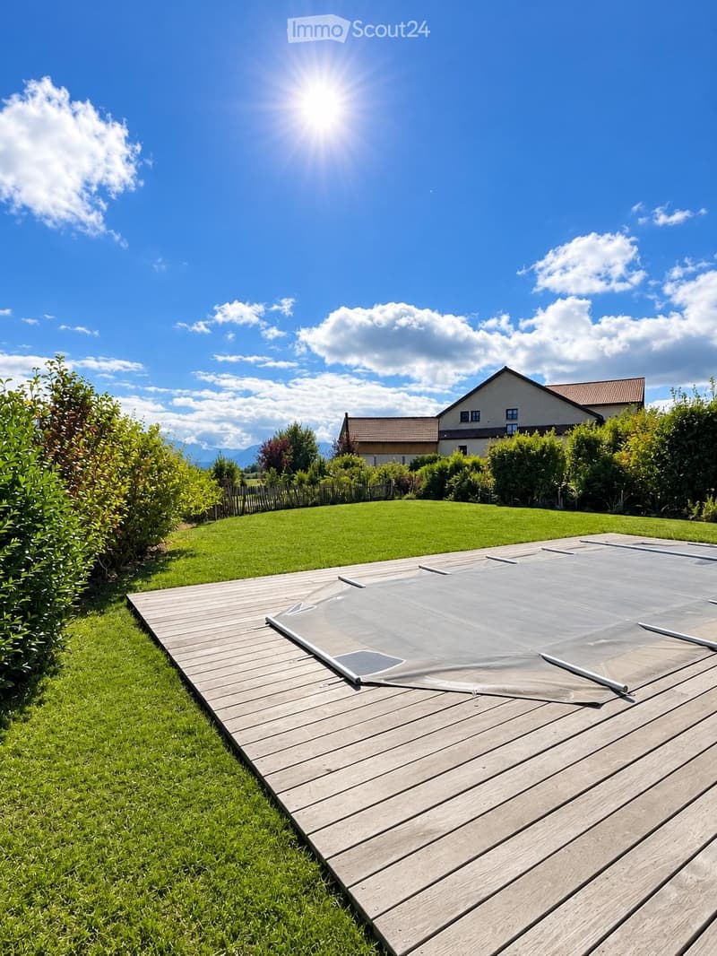 A wooden deck with a covered swimming pool, surrounded by green grass and bushes, with a house and mountains in the background.