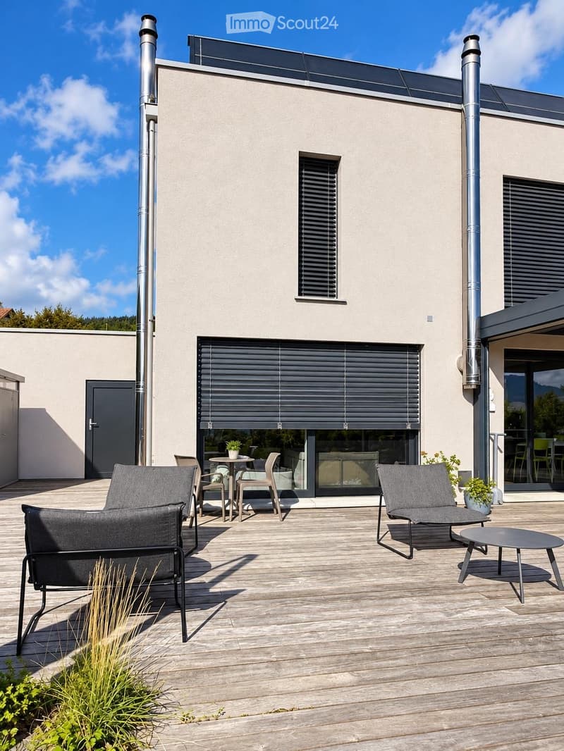 Modern wooden deck with grey outdoor furniture including a sofa, chairs, and a small table, with potted plants and a view of the house exterior.