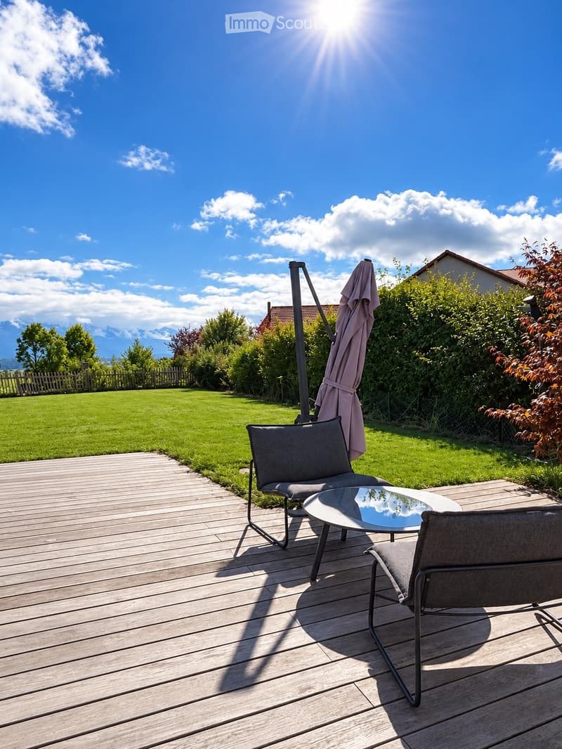 Wooden deck with two grey chairs and a round glass table, umbrella folded nearby, surrounded by a well-maintained lawn and greenery.