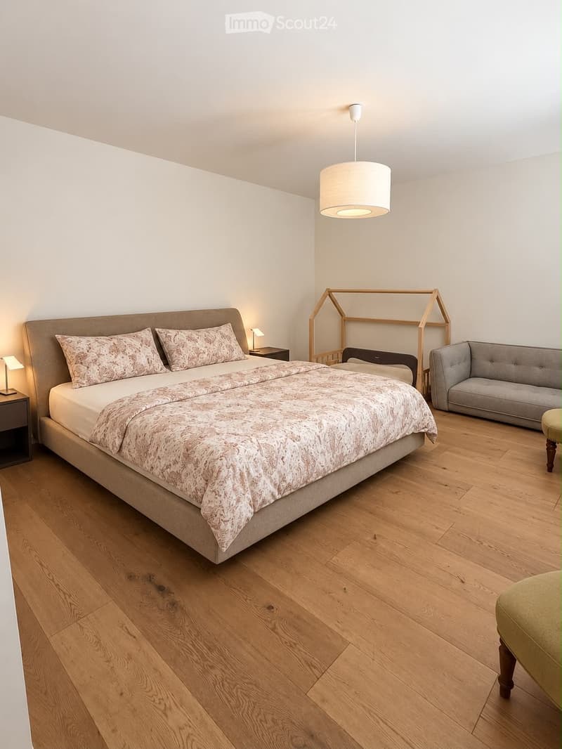 Modern bedroom with a large bed, patterned bedding, a grey sofa, a wooden stool, and a nightstand with a lamp. The room features light wooden flooring and white walls.