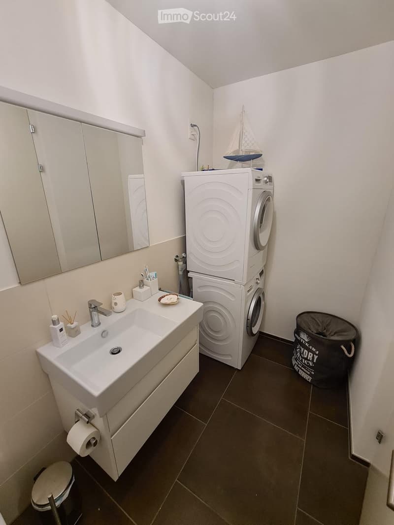 Modern bathroom with a white sink, mirror, toilet, and a stacked washing machine and dryer. Includes toiletries, a trash bin, and a small decorative model sailboat on top of the dryer.