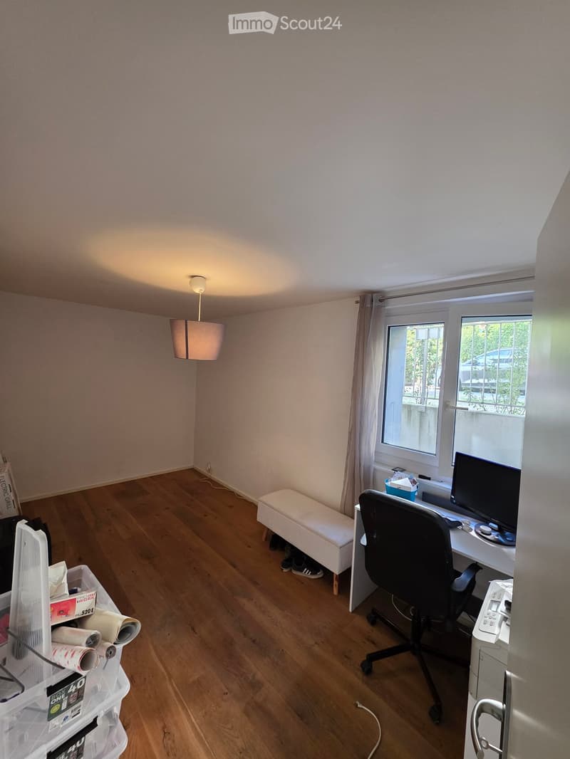 A furnished room with a wooden floor, a desk with a chair, a small white stool, a hanging lamp, and a window with a curtain. There is also a storage bin with rolls of paper towels on the left side.