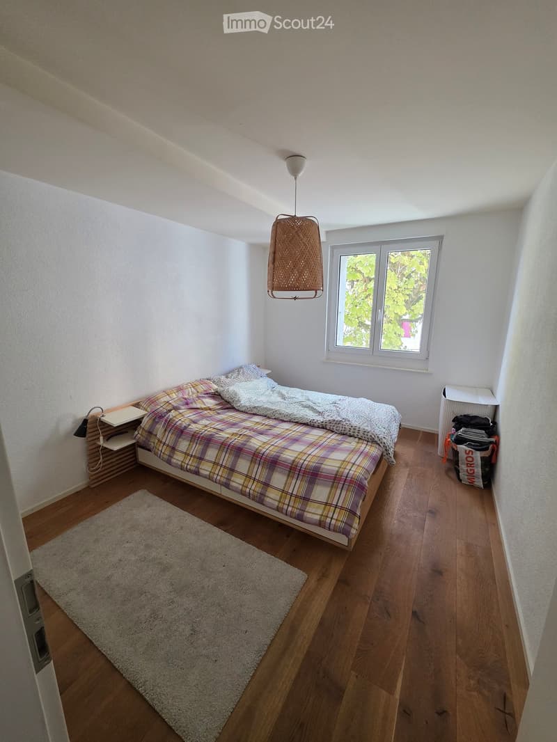 Cozy bedroom with wooden flooring, a single bed with plaid bedding, a nightstand with a lamp, and a small rug. A window with a view of greenery is visible.