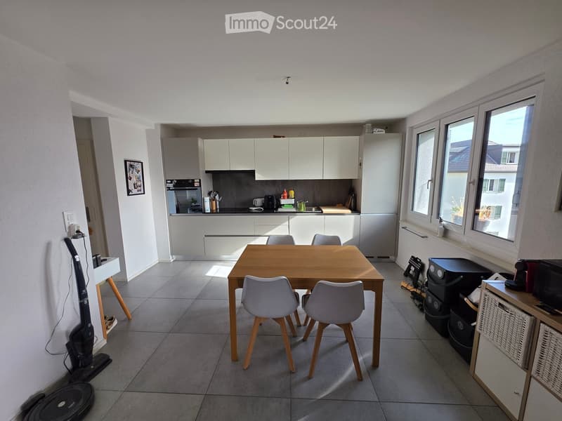 Modern kitchen with white cabinets, a wooden dining table with four chairs, and various appliances including a refrigerator, oven, and microwave. The space is well-lit with large windows and tiled flooring.