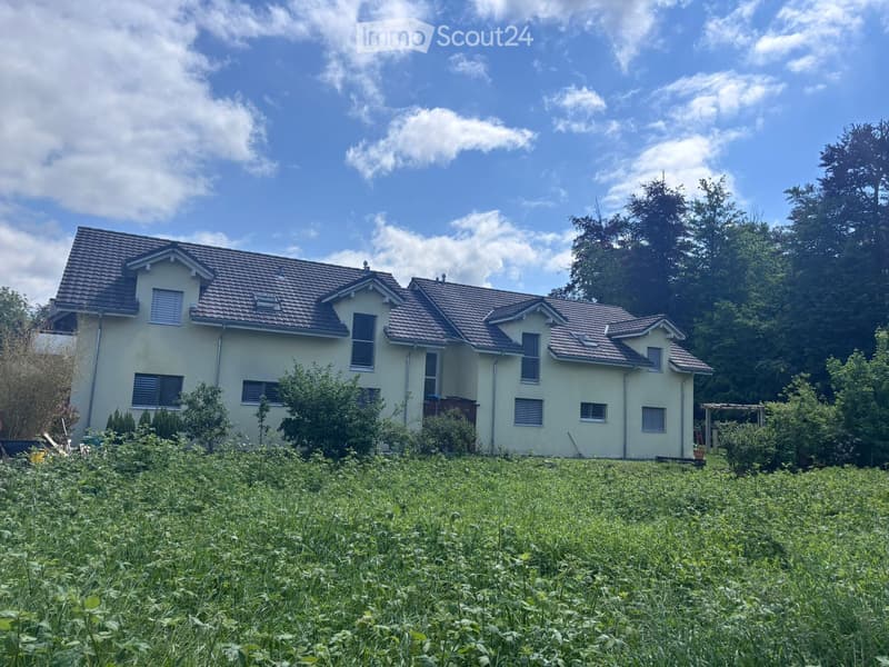 Two-story house with beige walls, brown tiled roof, multiple windows, surrounded by greenery and a grassy field.