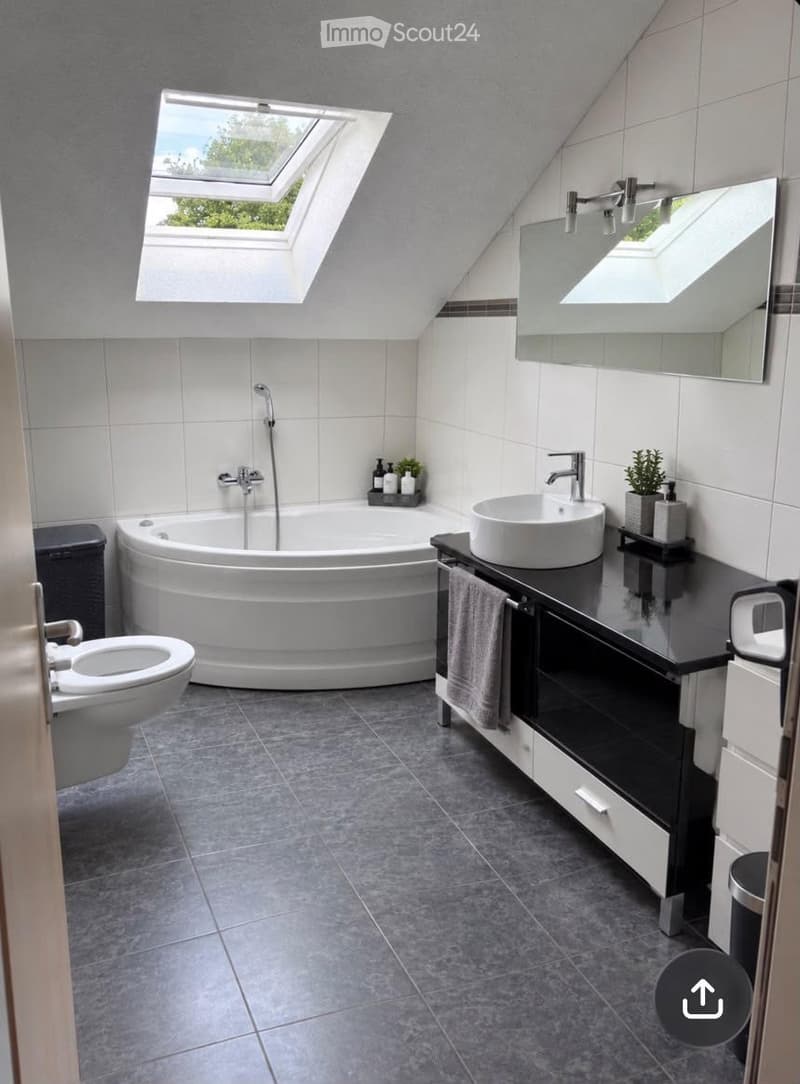 Modern bathroom with a large bathtub, toilet, sink, and a large mirror. The room features grey tiles and a skylight window above the bathtub.