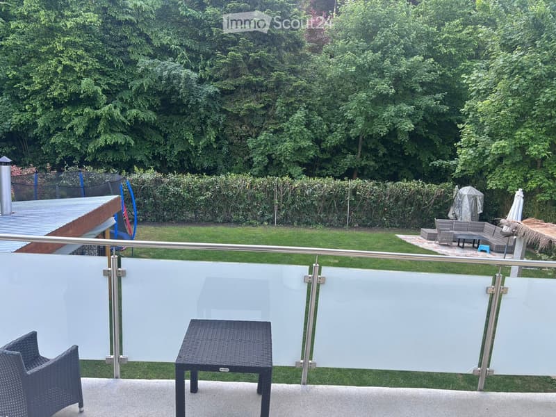 Balcony with a modern glass railing, featuring a wicker chair and a small table, overlooking a well-maintained garden with seating area and umbrella.
