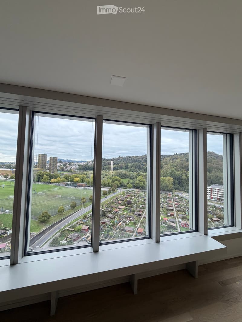 Empty room with large windows offering a view of a neighborhood, a soccer field, and surrounding greenery. A white bench is positioned beneath the windows.