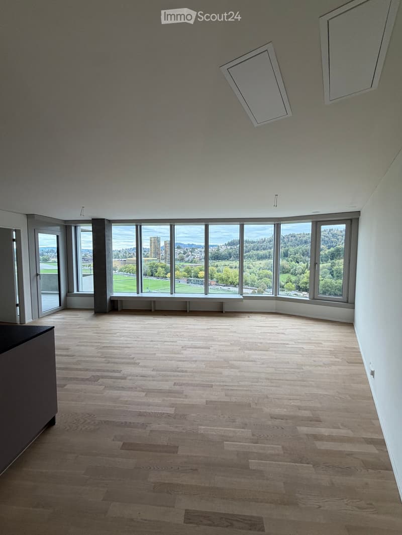 Empty room with wooden flooring, large windows offering a view of a park and cityscape, and a built-in bench along the window wall.