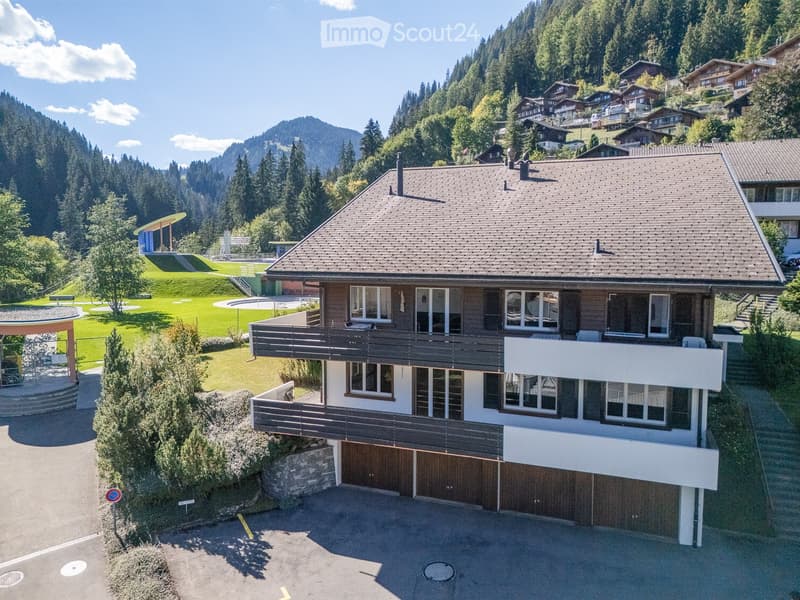 A two-story house with a tiled roof, large windows, and a wooden balcony. The house features a garage and is surrounded by a well-maintained lawn and trees, with a scenic mountain backdrop.