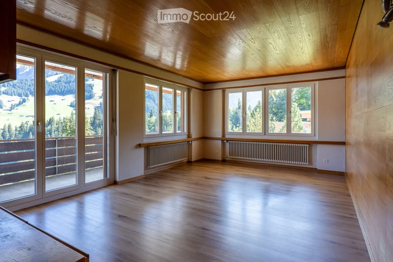 Empty room with wooden floors, large windows offering a scenic mountain view, and radiators along the walls.