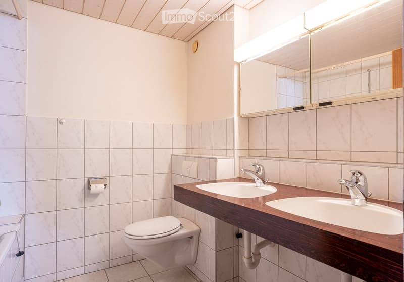 Modern bathroom with white tiled walls and floor, double sink vanity with wooden countertop, chrome faucets, toilet, and large mirror above the sinks.