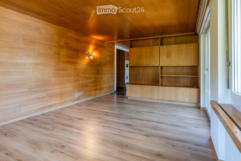 Empty hallway with wooden flooring and walls, built-in wooden shelving unit, and a recessed light fixture on the wall.