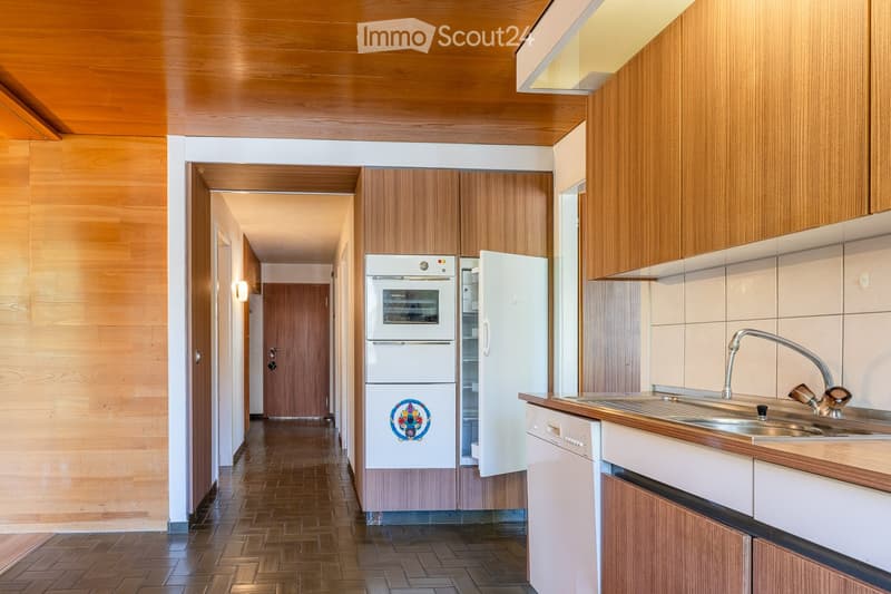 Modern kitchen with wooden cabinets, stainless steel sink, refrigerator, oven, and dishwasher. The space features a wooden floor and warm lighting.