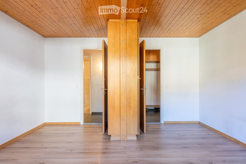 Empty room with light wooden flooring, white walls, and a large wooden wardrobe. The room has a wooden ceiling and visible doorways leading to other spaces.