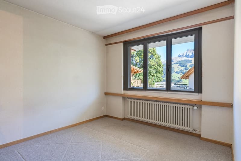 Empty room with beige walls, carpeted floor, large window offering a scenic mountain view, radiator below the window, and wooden trim around the window and baseboards.