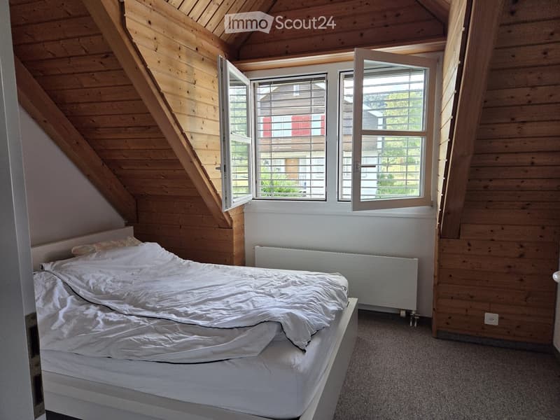Cozy attic bedroom with wooden walls, slanted ceiling, large windows, and a neatly made bed.