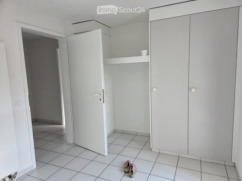 Empty room with white tiled floor, white walls, built-in closet, and a small open doorway leading to another space. A pair of shoes is placed on the floor.