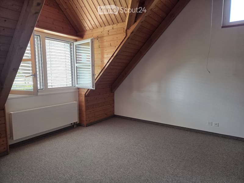Empty attic room with wooden slanted ceilings, large windows with blinds, carpeted floor, radiator, and electrical outlets.