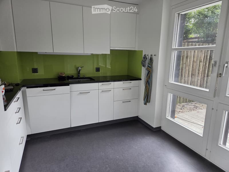 Modern kitchen with white cabinets, black countertops, and a green backsplash. Features a sink, faucet, and a towel hanging on the wall. A large window offers a view of the outdoor area.