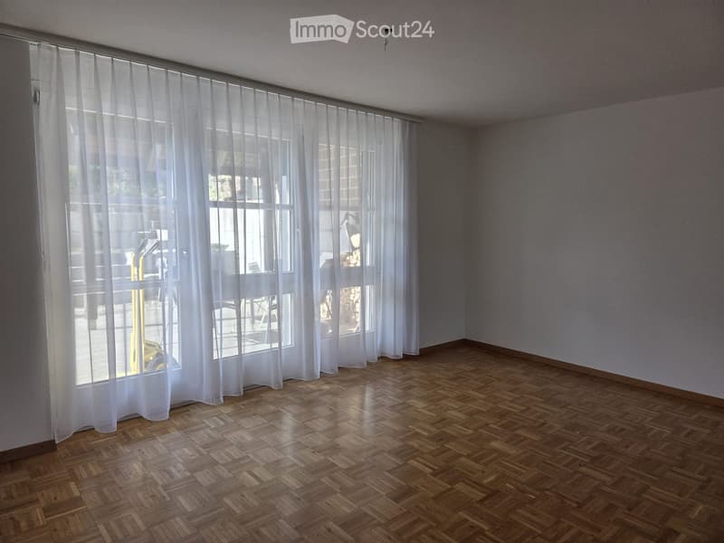 Empty room with wooden herringbone flooring, large windows with white curtains, and neutral-colored walls.
