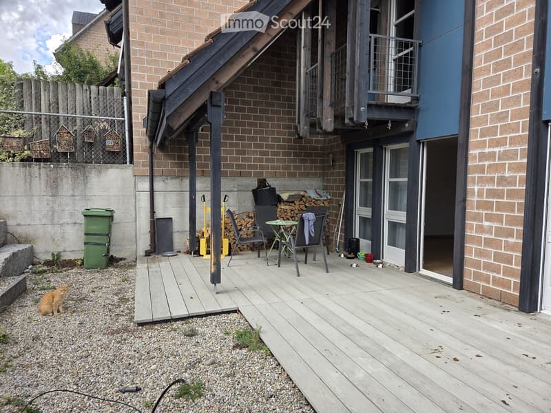 Wooden deck area with a small table, chairs, a cat, and a stack of firewood near the entrance of a building.