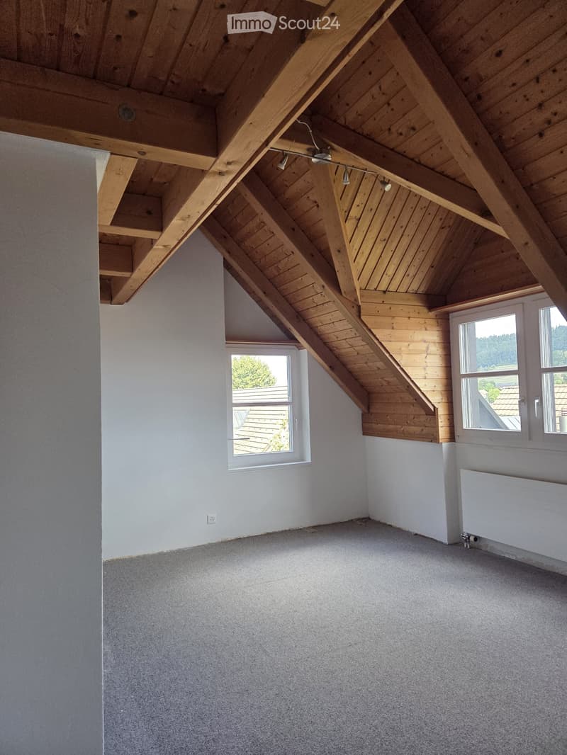Empty attic room with wooden slanted ceilings, grey carpet, white walls, and windows letting in natural light.