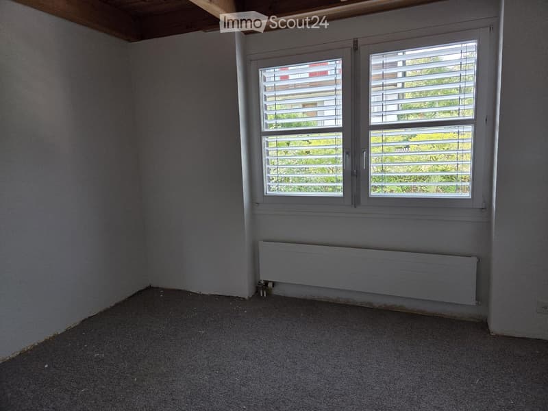Empty room with grey walls, carpeted floor, and a radiator under a window with blinds.
